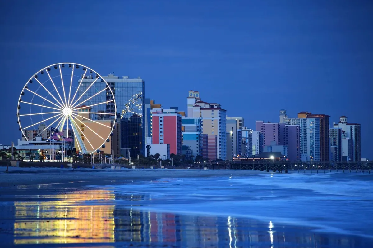 A vibrant exterior scene featuring a large Ferris wheel, colorful beachfront buildings, and gentle ocean waves. The image captures a lively amusement area that is bustling with activity, with the Ferris wheel offering sweeping views over the beach and sunset-lit sky.