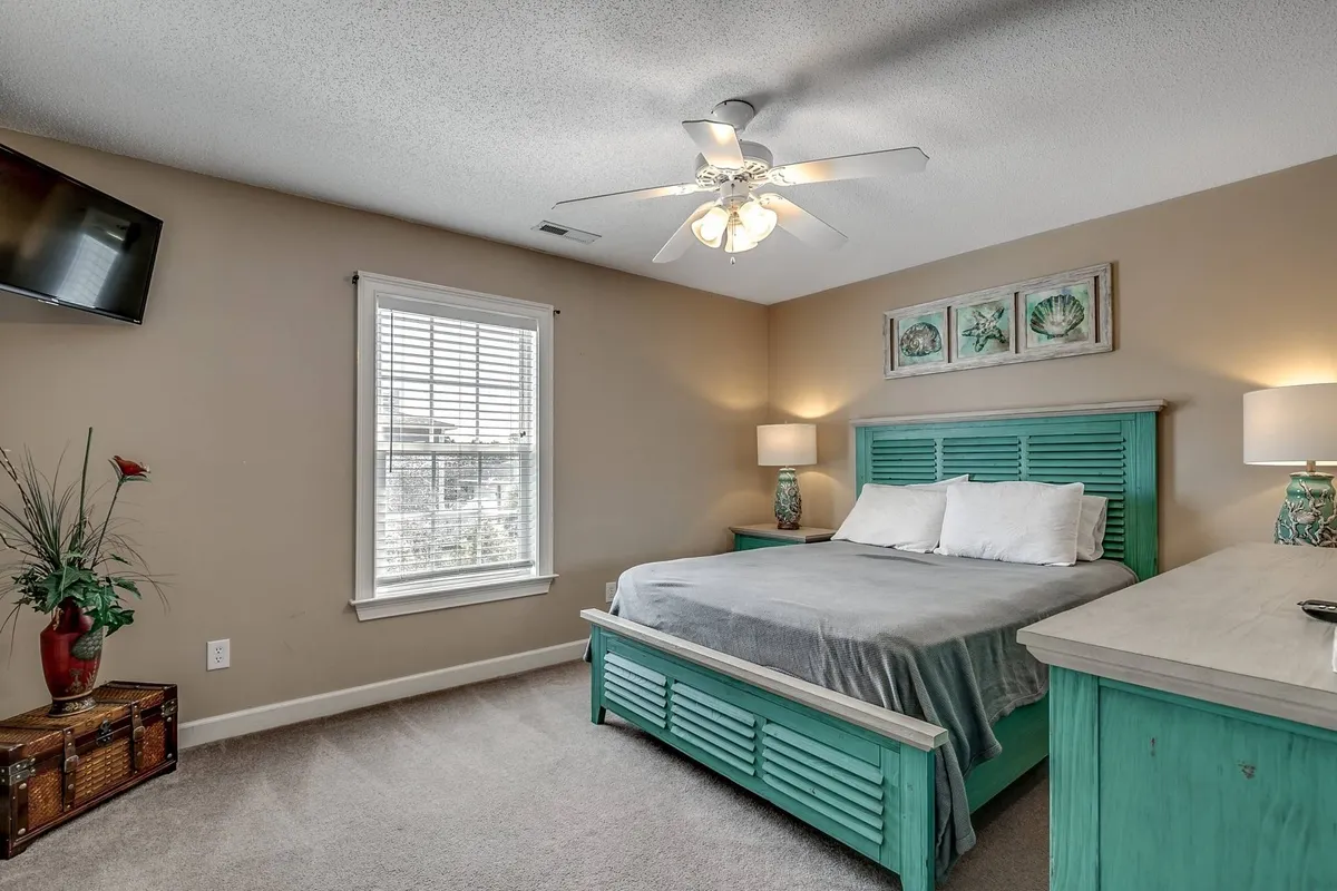 A serene bedroom with a turquoise bed frame, white bedding, decorative wall art, a ceiling fan, and a TV. Natural light filters in, creating a tranquil space ideal for resting or enjoying a private movie night.