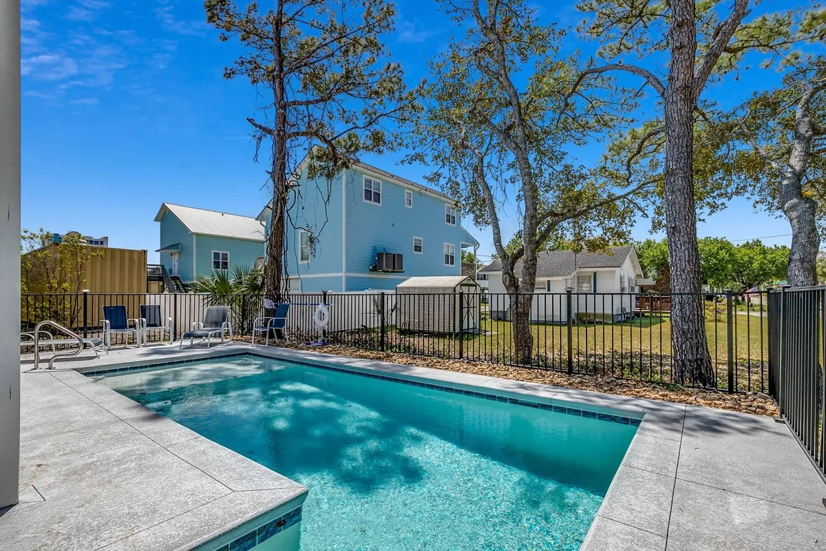 This exterior image showcases a sparkling swimming pool surrounded by lush trees, with a blue house and fence providing a sense of privacy and relaxation. The open sky and clear water suggest an inviting space perfect for both sunbathing and social gatherings.