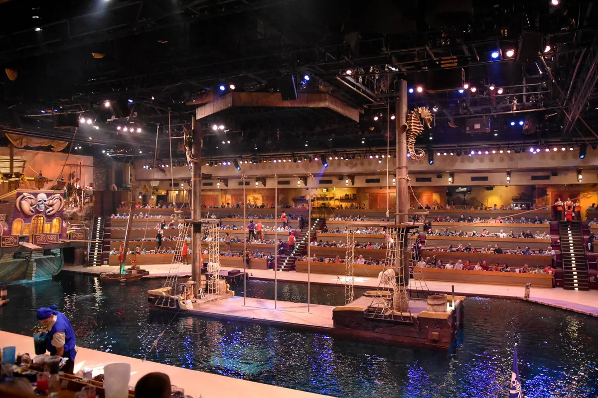 An interior scene showcasing a dynamic stage set with aquatic features and dramatic ship props. The audience seating is filled, and the mood is lively, reflecting the thrill of a live performance and an immersive dining experience.