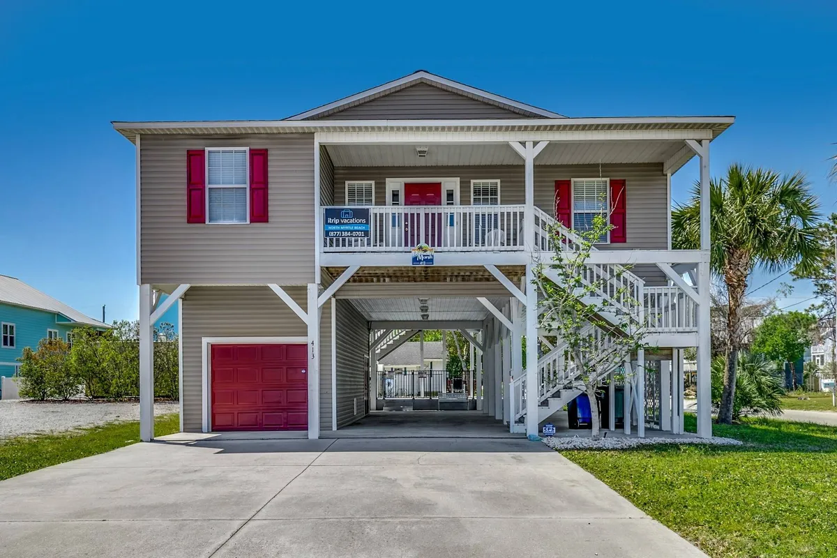 The image showcases the cheerful exterior of a single-family house in North Myrtle Beach, highlighted by bright red doors, inviting stairs leading up to the entrance, and lush palm trees swaying in the background. The spacious garage and welcoming porch set the scene for family fun and relaxation, evoking visions of sunset views and friendly gatherings in the yard.