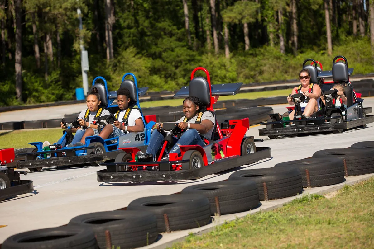 An energetic outdoor go-kart track with racers zipping around curves, tire barriers, and a sense of action and excitement in the air. Families and friends are enjoying the thrill of friendly competition and laughter under the open sky.