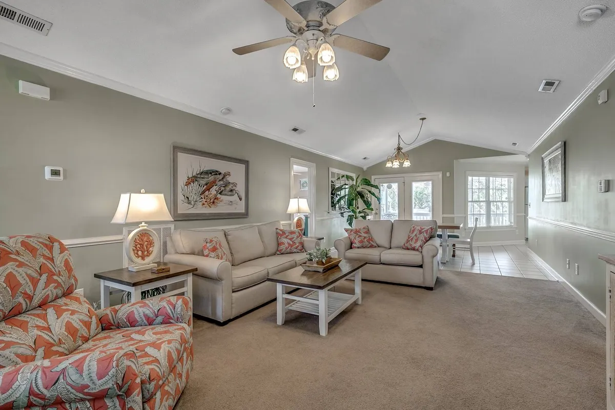 A bright and welcoming living area featuring a comfortable seating arrangement, decorative artwork on the walls, a leafy plant by the window, and a stylish lighting fixture overhead. The space radiates warmth and is perfect for gathering with friends or relaxing quietly.